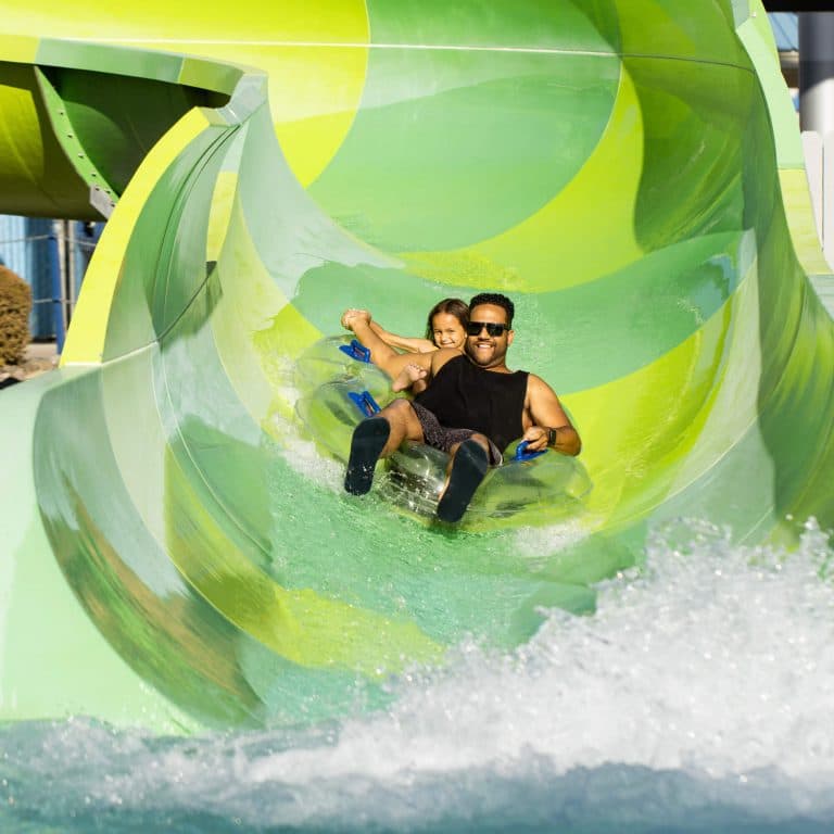 A man and a child joyfully slide down a twisting, green water slide in a clear inflatable tube, splashing into the water below on a sunny day.