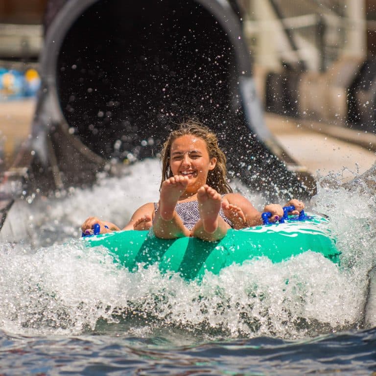 A person joyfully emerges from a waterslide, riding a green inflatable tube. Water splashes around, capturing a moment of excitement and fun at the Waterpark.