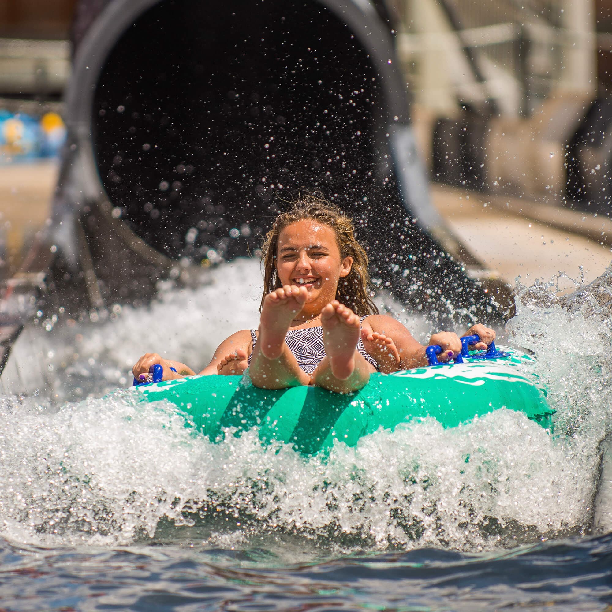 A person joyfully emerges from a waterslide, riding a green inflatable tube. Water splashes around, capturing a moment of excitement and fun at the Waterpark.